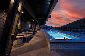 Ein Poolbereich im Freien mit Blick auf den Sonnenuntergang, eine überdachte Lounge mit Sofas und Tischen sowie eine Backsteinmauer.