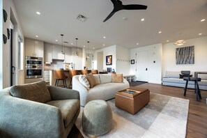 Ein modernes Wohnzimmer mit einer Couch, einem Hocker und einem Couchtisch, eine Küche mit Barhocker und ein Essbereich mit Tisch und Stühlen.