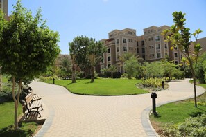 A landscaped courtyard with a curved pathway, benches, and a building in the background. - SODIC Kattameya Plaza, 3-Bedroom, 230 m² Family Apartment, 3 Pools in New Cairo (New Cairo)