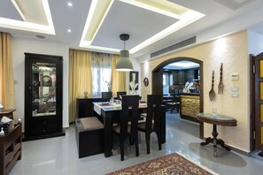 A modern dining area with a chandelier, a dining table set for four, and a kitchen visible through an archway. - SODIC Kattameya Plaza, 3-Bedroom, 230 m² Family Apartment, 3 Pools in New Cairo (New Cairo)