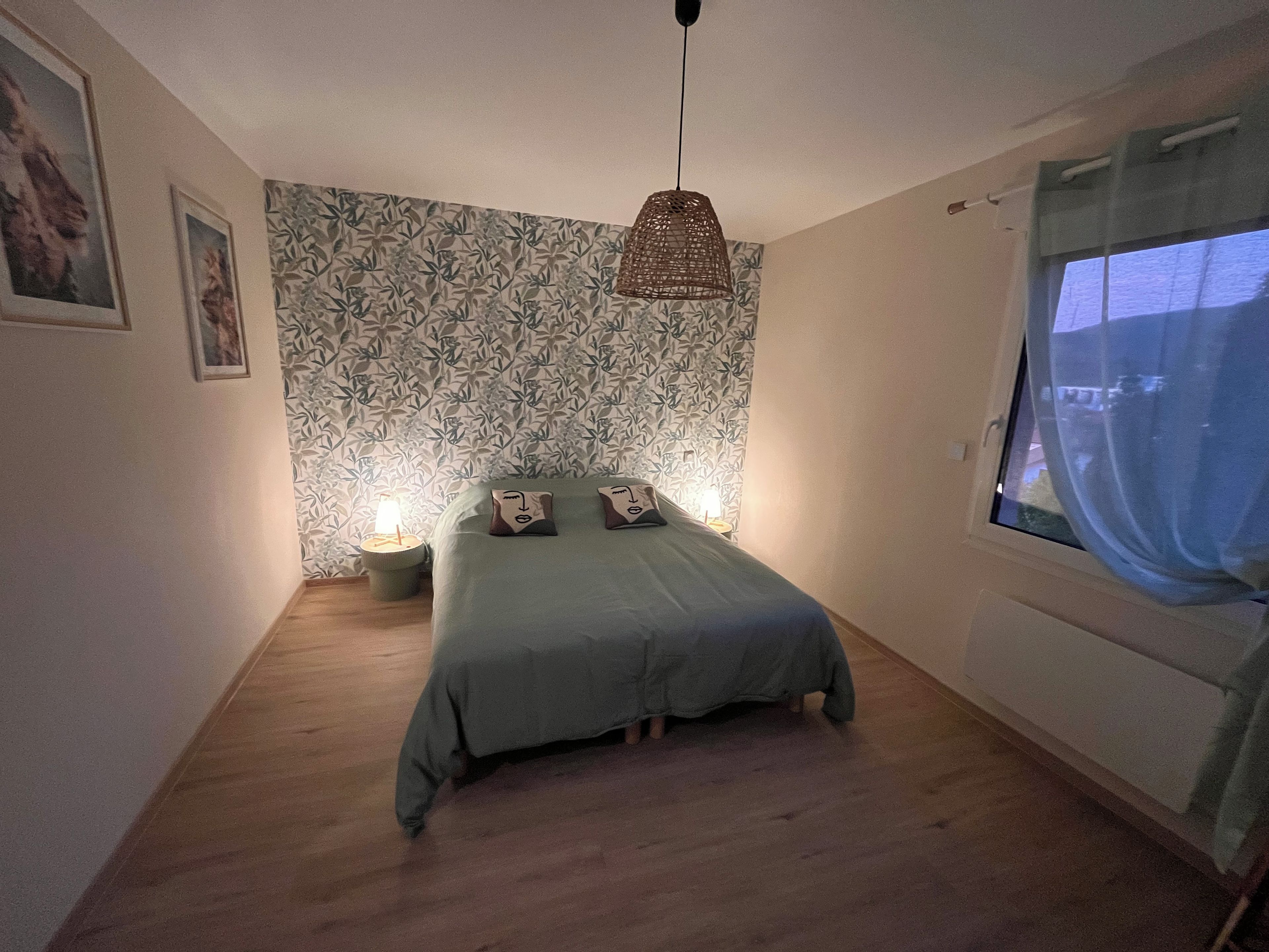 A bedroom with a bed, two bedside lamps, a window with curtains, and a patterned wallpaper.