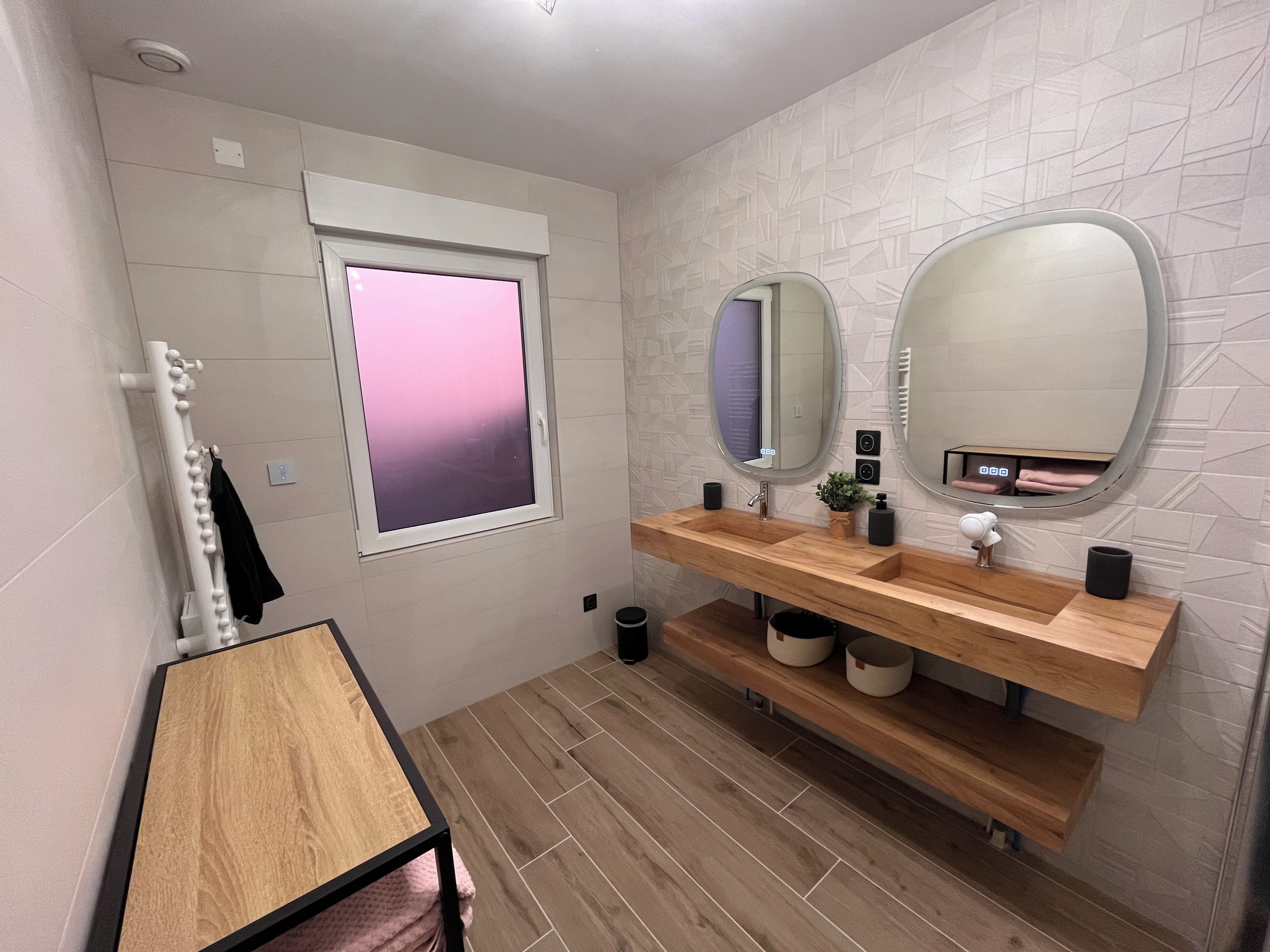 Ein modernes Badezimmer mit einem Holzbadschrank, zwei Spiegeln, einem Fenster mit Blick auf Rosa und einem Handtuchhalter.
