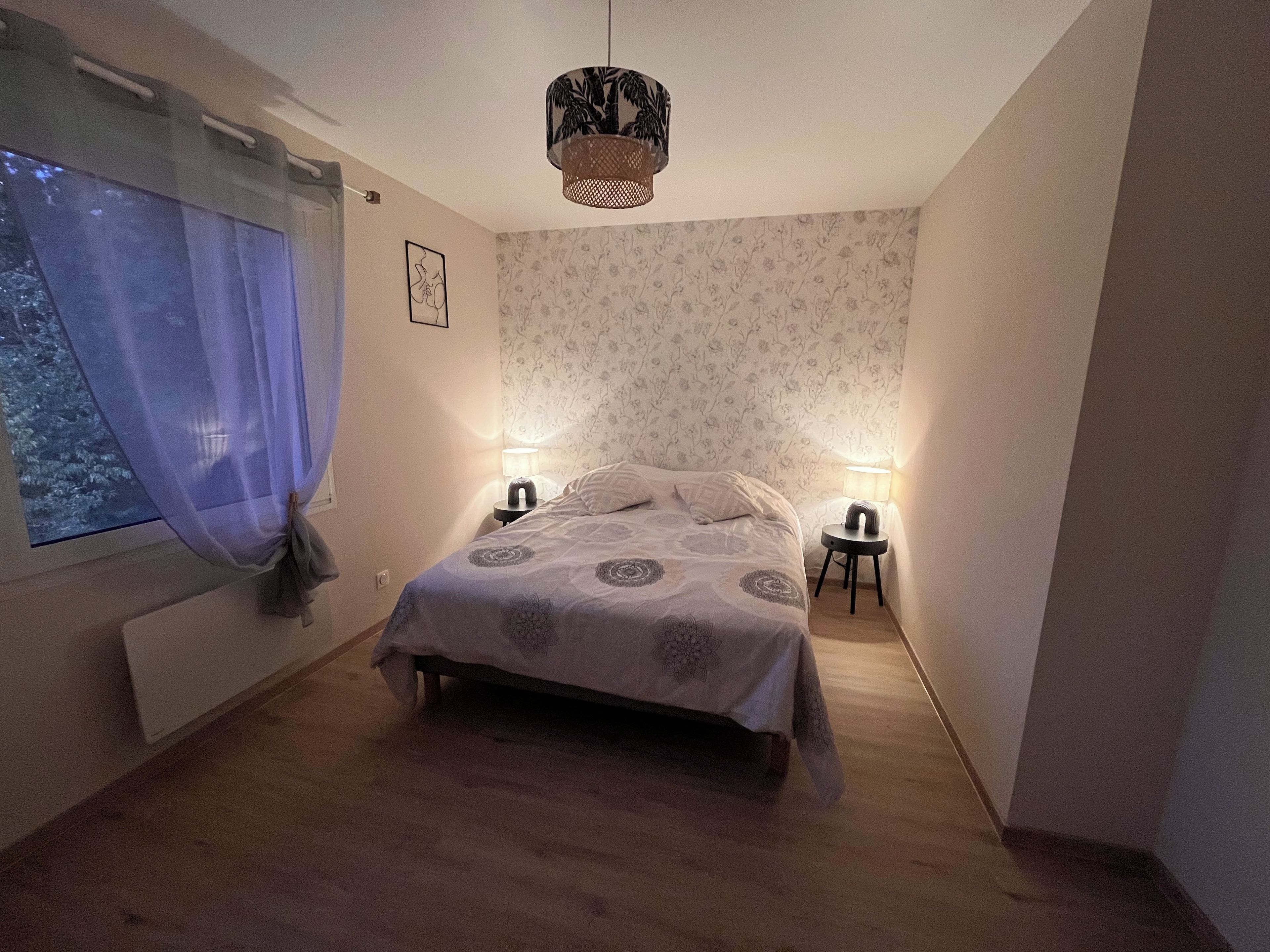Une chambre à coucher comprenant un lit, deux tables de chevet, une fenêtre avec des rideaux, une lampe fixée au mur et un luminaire suspendu.