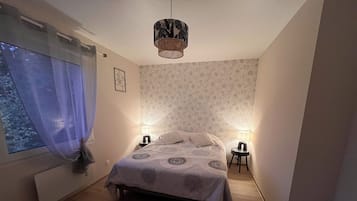 Une chambre à coucher comprenant un lit, deux tables de chevet, une fenêtre avec des rideaux, une lampe fixée au mur et un luminaire suspendu.
