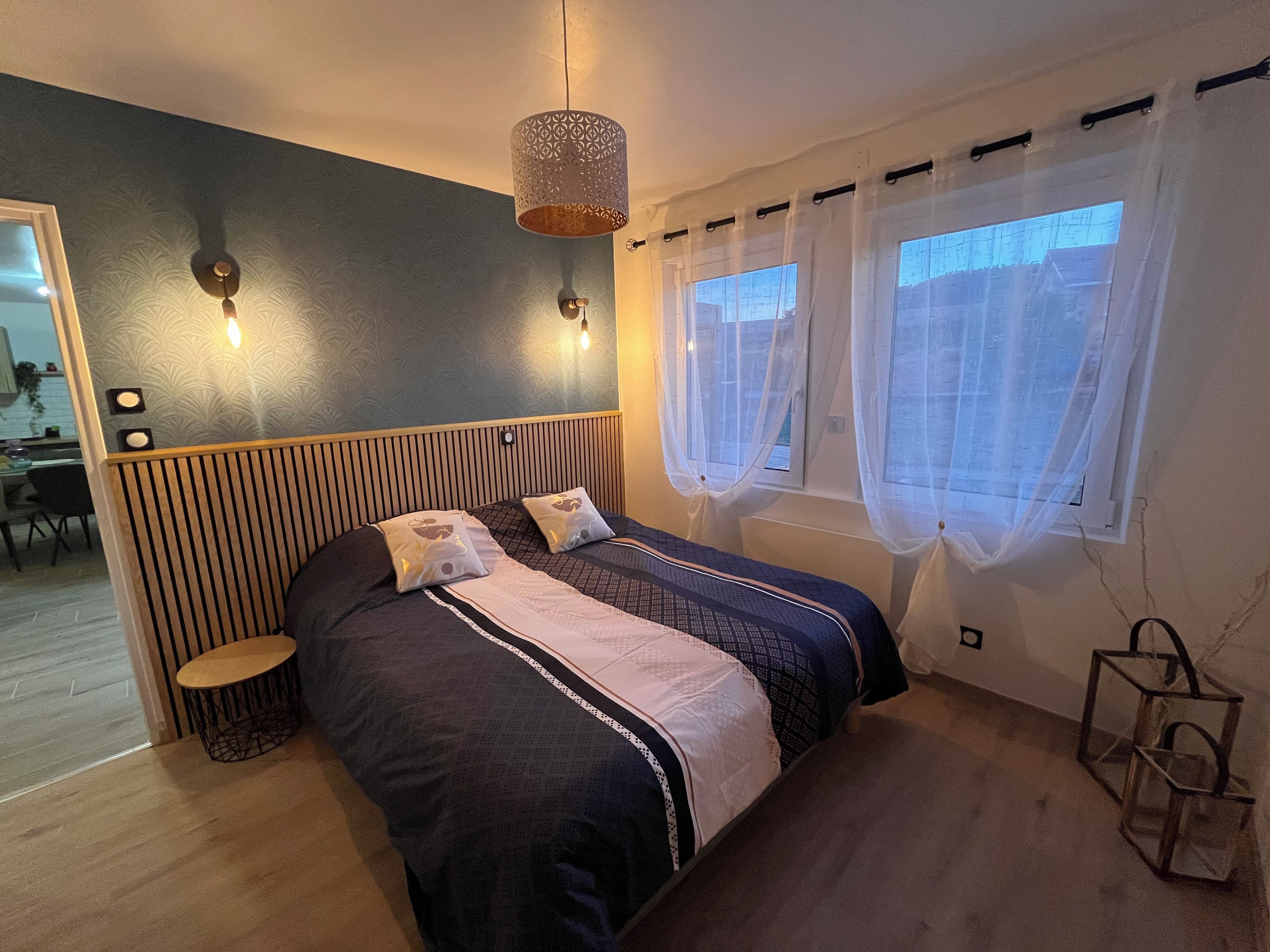 A bedroom with a bed, bedside tables, a hanging light fixture, a window with sheer curtains, and a view of the outside.