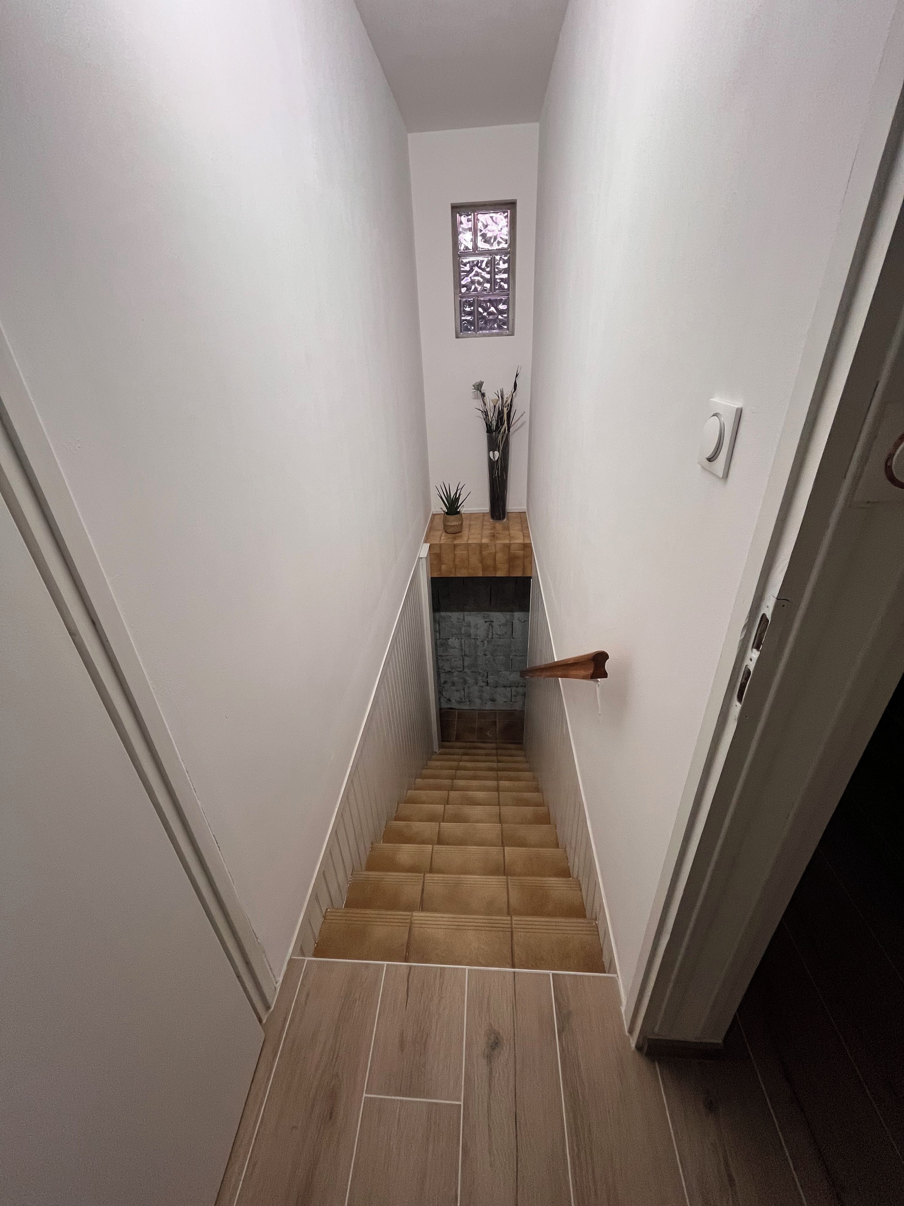 A narrow staircase with wooden steps and white walls, leading to a landing with a small window and a plant.