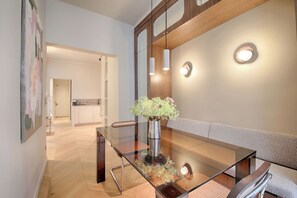 A modern interior with a glass dining table, a vase with flowers, a sofa, and a kitchen in the background. - Louvre Palais Royal New Luxury Flat (Paris)
