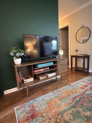 Een woonkamer met een flatscreen televisie op een houten standaard, een klein tafeltje met een spiegel, en een kleurrijk tapijt op de vloer.