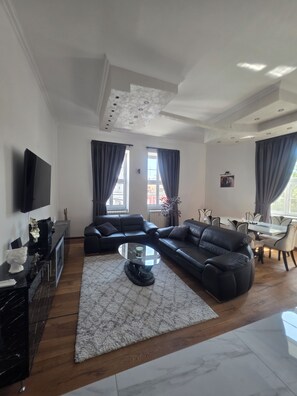 Een moderne woonkamer met een hoekbank, een flatscreen televisie, een eethoek met tafel en stoelen, en grote ramen met gordijnen.