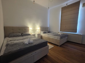 A bedroom with a large bed, two bedside tables, a window with blinds, and a radiator. - Elegant Historic Apartment in the Heart of Vršac! (Vršac)