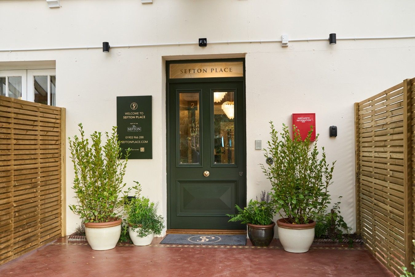 Una puerta verde con letrero 'SEFTON PLACE', un cartel de bienvenida e información sobre seguridad contra incendios.