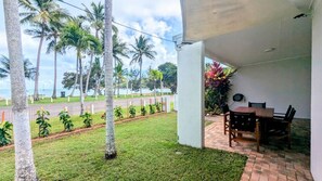 Un patio techado con una mesa y sillas, con vistas a una zona verde con palmeras y al mar.