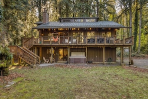 A two-story wooden cabin with a balcony, a hot tub, and a covered porch area. - River Hutte – Hot Tub, Pool Table & River Front (Mount Hood Village)
