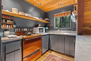 Eine moderne Küche mit Einbauofen, Mikrowelle und Spüle, Holzregalen und einem Fenster mit Blick auf Bäume.