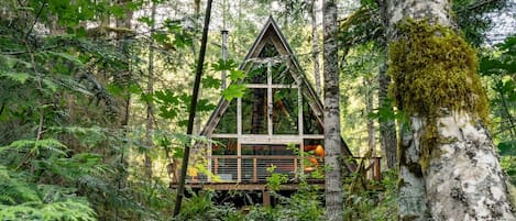 A cabin nestled in a forest, with a triangular roof and wooden deck.