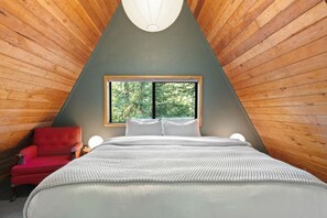 Ein Schlafzimmer mit schrägen Wänden, einem Bett mit weißer Bettwäsche, einem roten Sessel und einem Fenster mit Blick auf Bäume.