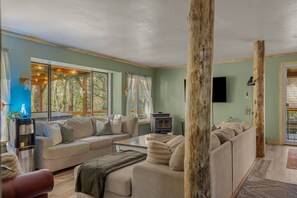 A living room with a sofa, coffee table, and a fireplace. There are large windows and a view of trees outside. - Mahalo Mountain Cabin *Games, Hot Tub & Fire Pit!* (Brightwood)