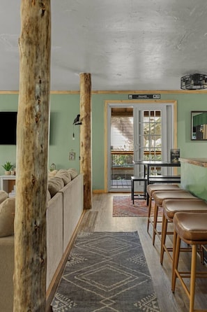 A cozy living area with a sofa, wooden beams, a dining table with stools, and a view through a window. - Mahalo Mountain Cabin *Games, Hot Tub & Fire Pit!* (Brightwood)
