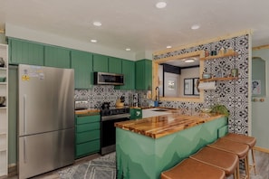 Une cuisine avec des meubles de couleur verte, un réfrigérateur en acier inoxydable, un four à micro-ondes et un îlot central en bois avec des tabourets.