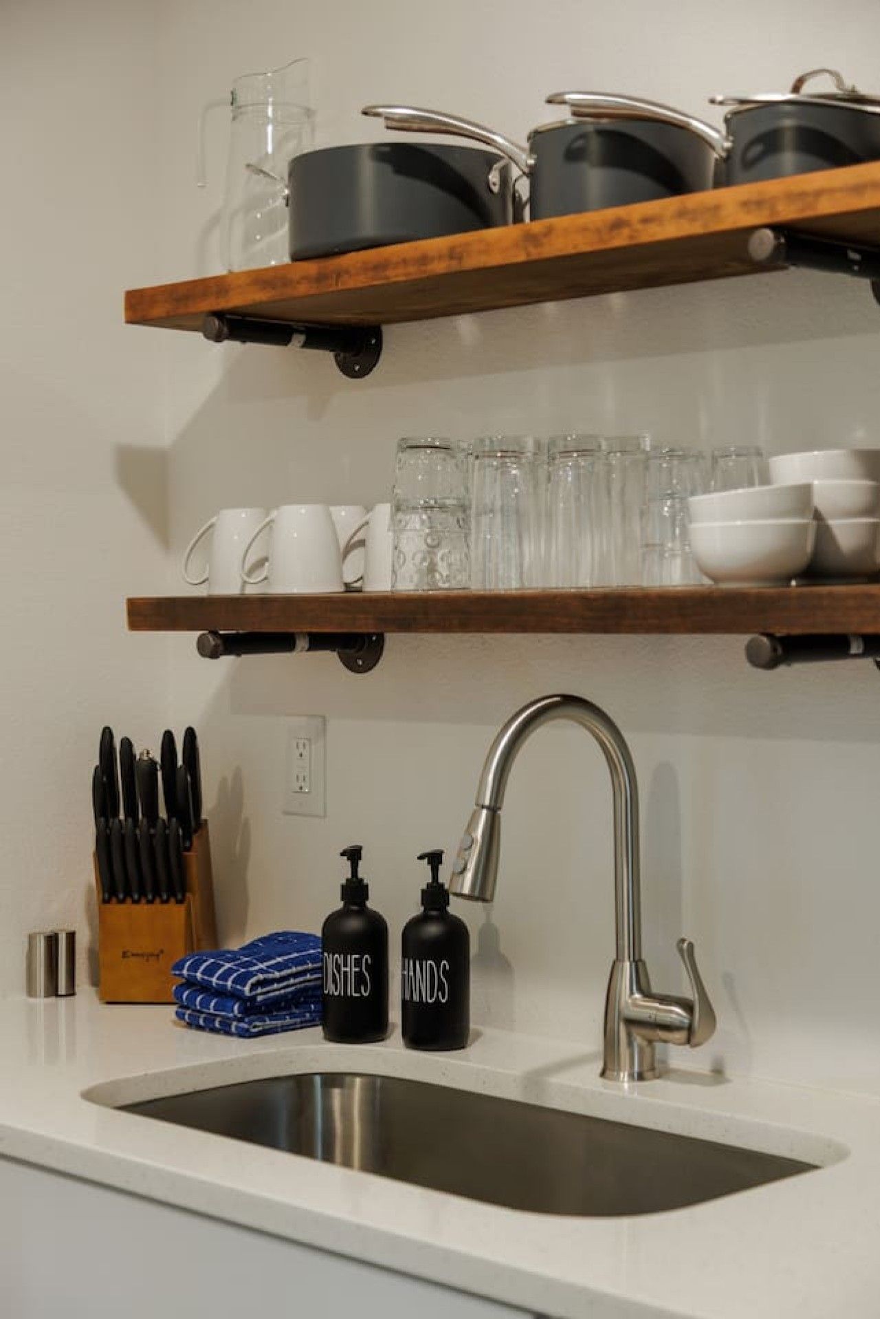 Una cocina con un lavamanos de acero inoxidable, repisas de madera con cristalería y utensilios de cocina, y un bloque para cuchillos con cubiertos.