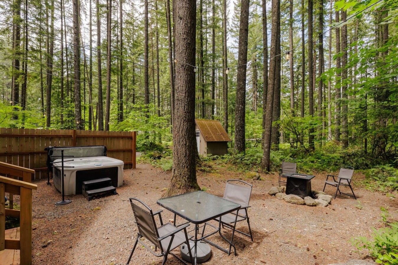A forested outdoor area with a hot tub, a table with chairs, and a small wooden structure.