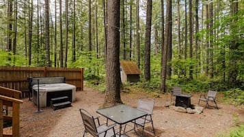 A forested outdoor area with a hot tub, a table with chairs, and a small wooden structure.