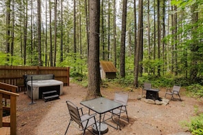 A forested outdoor area with a hot tub, a table with chairs, and a small wooden structure. - Mt Hood Hygge- A Cozy Tiny Cabin with Hot Tub (Mount Hood Village)
