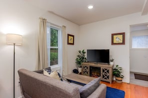 A living room with a sofa, a flat-screen TV on a wooden stand, a floor lamp, and a window with curtains.