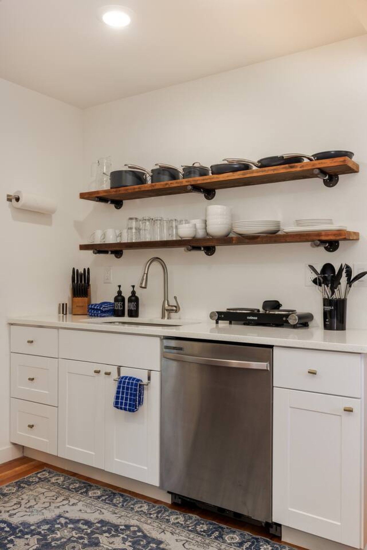 A kitchen with white cabinets, a stainless steel dishwasher, and wooden shelves above the sink holding dishes and cookware.