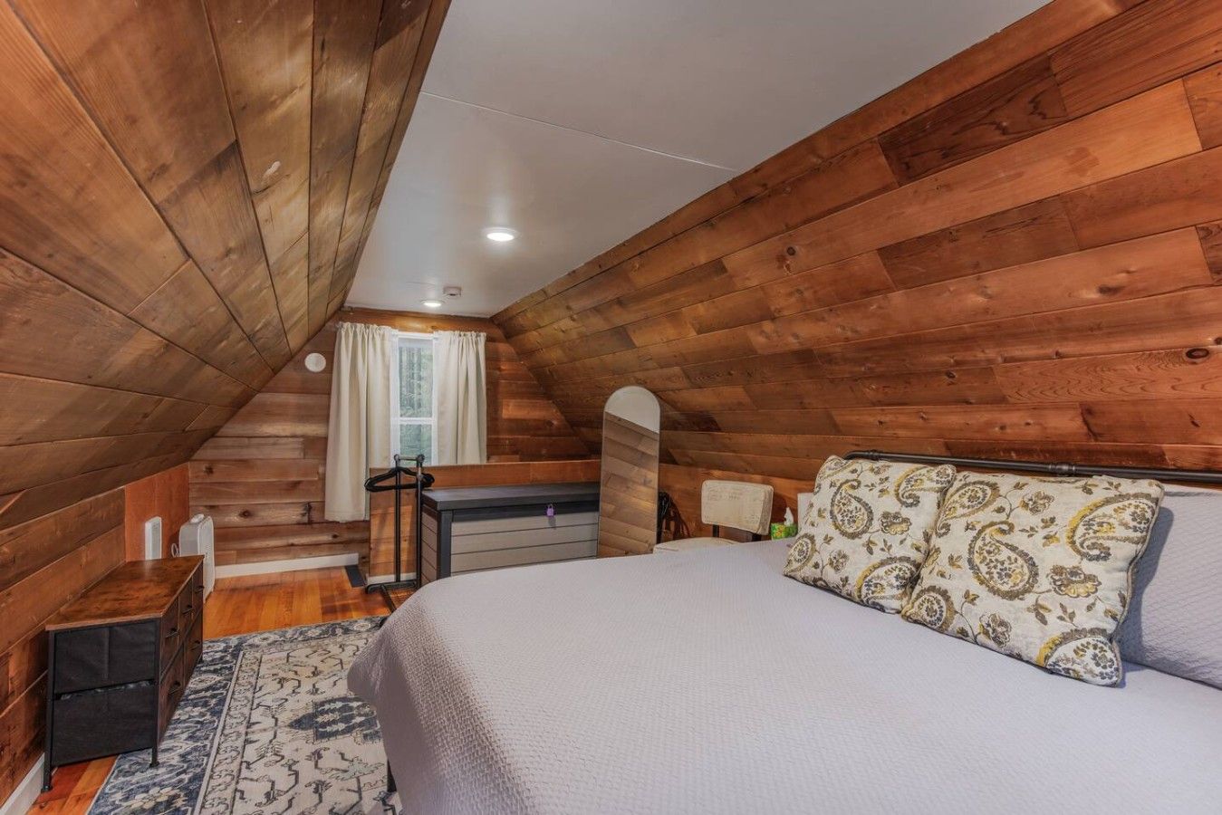 A cozy attic bedroom with wooden walls, a bed, a desk, and a window with curtains.