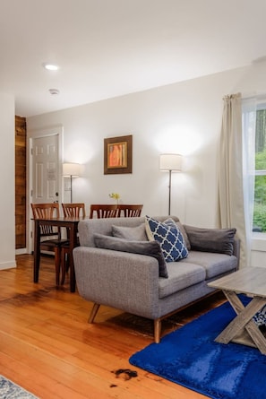 A living room with a grey sofa, wooden dining table, and chairs, a blue rug, and a painting on the wall.