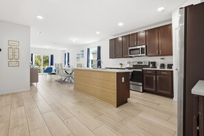 A modern kitchen with wooden cabinets, a central island, and a dining area with blue chairs. - Near Disney 4BR Family Home with Super Mario Room (Kissimmee)