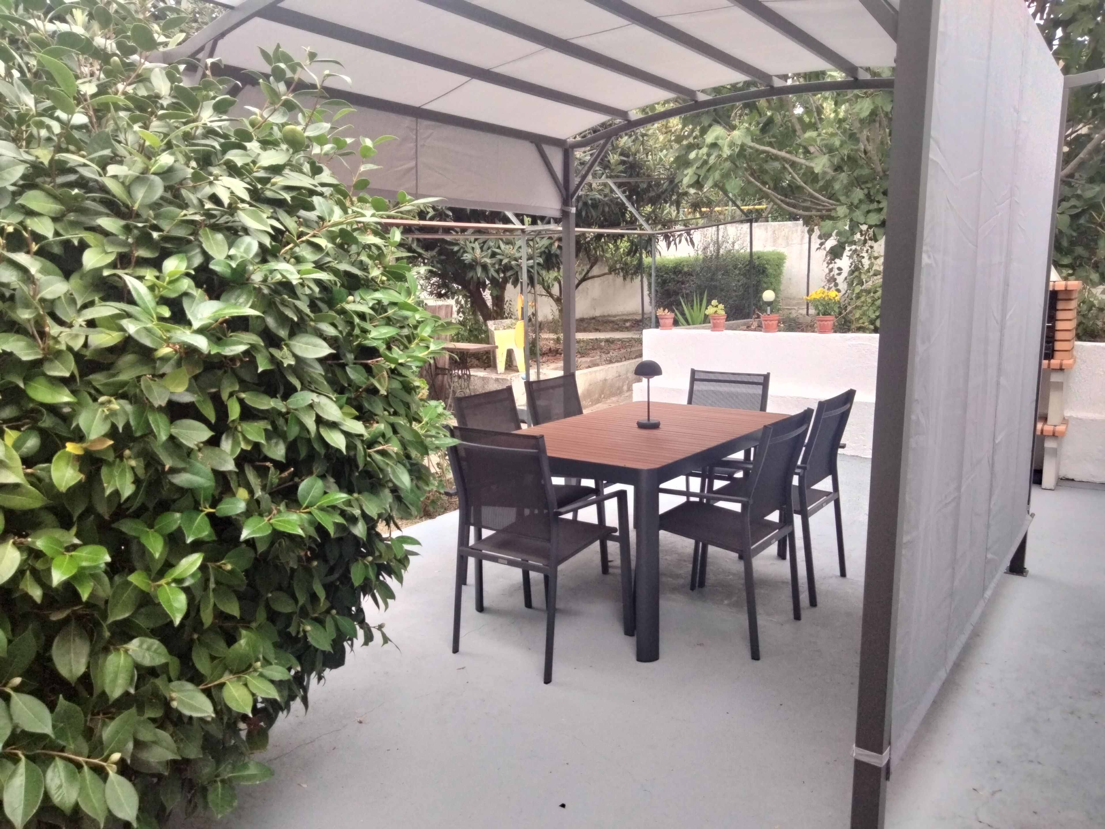An outdoor patio with a covered table and chairs, surrounded by greenery.