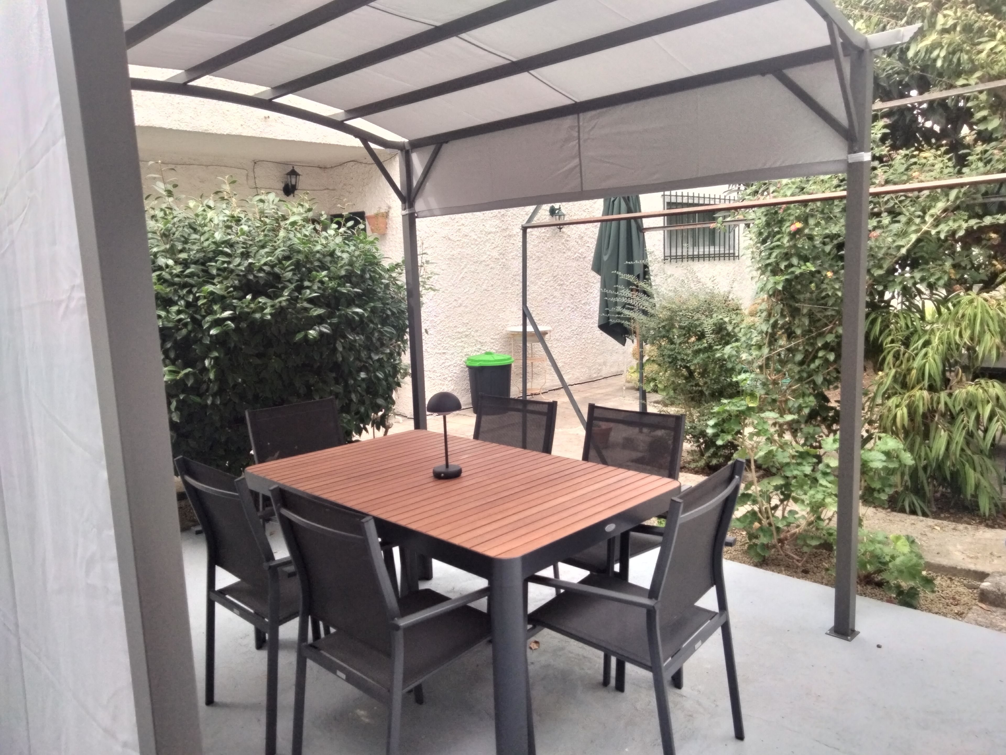 Eine Terrasse im Freien mit Tisch und Stühlen, umgeben von Pflanzen und einem überdachten Bereich.