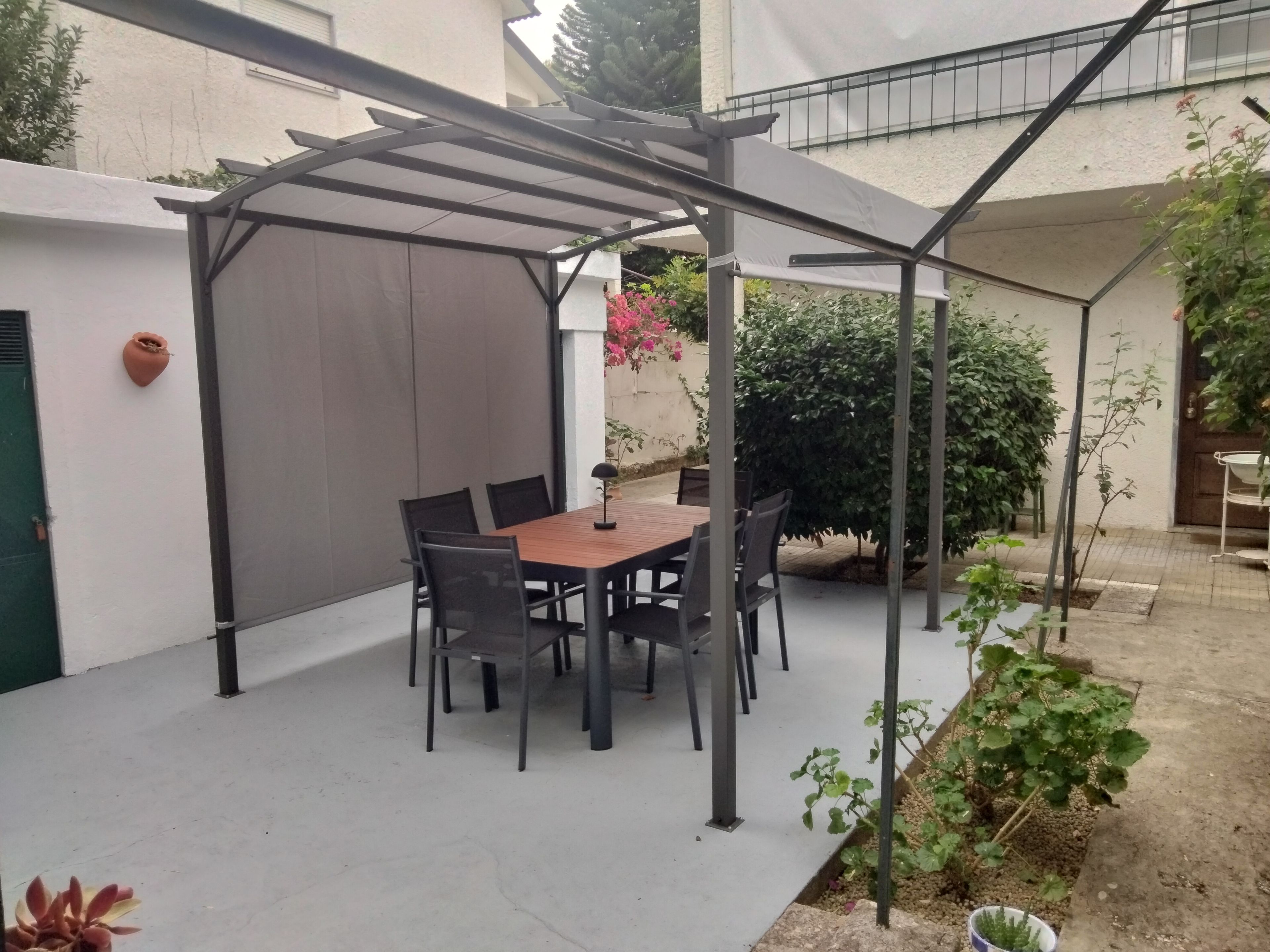 Een overdekt terras met een tafel en stoelen, omgeven door beplanting en een gebouw.
