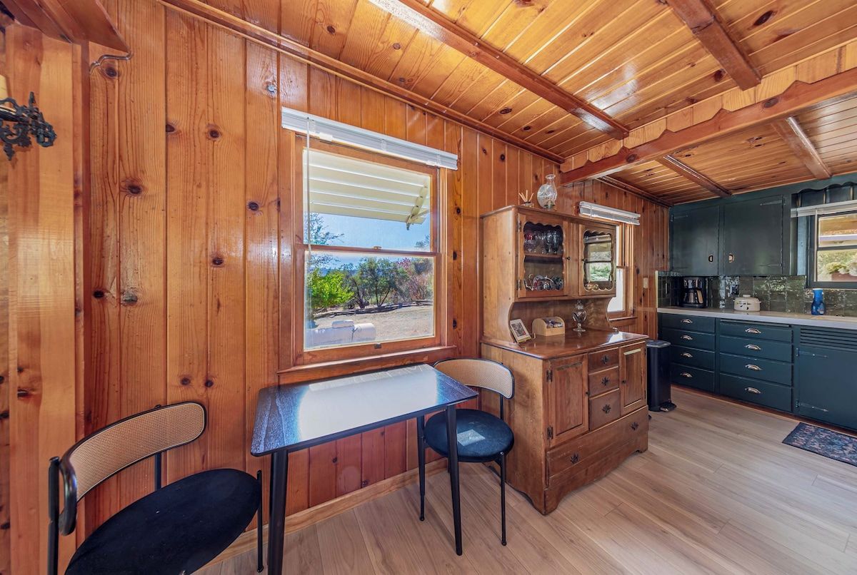 Un intérieur en bois comprenant une table à manger, des chaises, un coin cuisine et une fenêtre avec des stores.