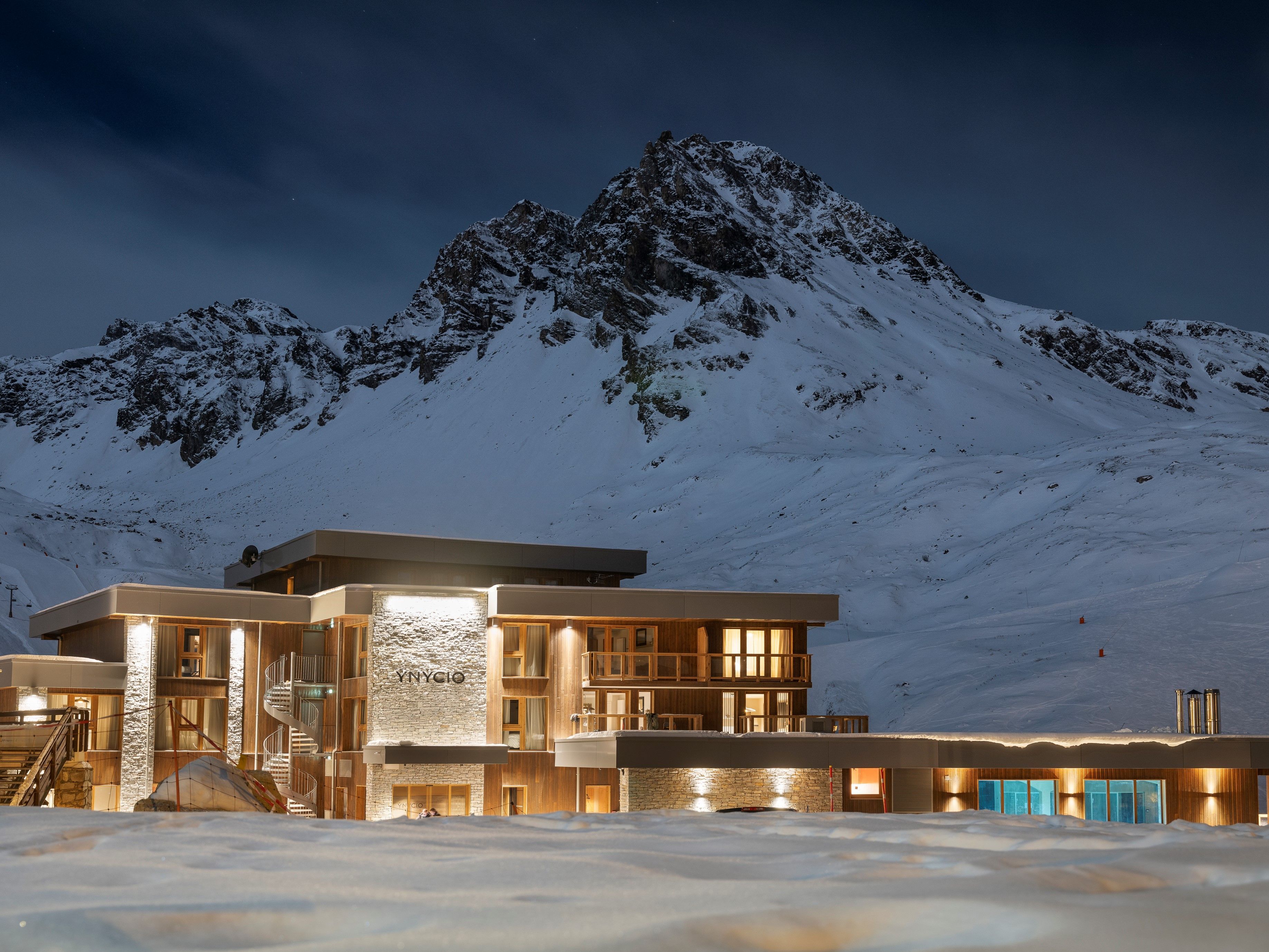 A modern hotel with a stone facade, multiple balconies, and a view of a snow-covered mountain.