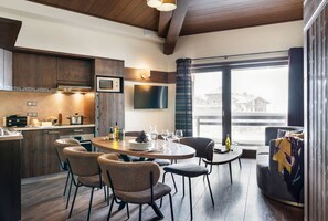A modern kitchen with wooden cabinetry, a dining area with a round table and chairs, and a large window offering a view of buildings. - Au pied des pistes | Piscine | Esprit famille (Val Thorens)