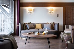 A modern living room with a grey sofa, two wall-mounted lamps, a coffee table with a book, a glass of milk, and a plate with bread and an apple. - Skis aux pieds | Piscine & spa | Vue montagne (Val Thorens)