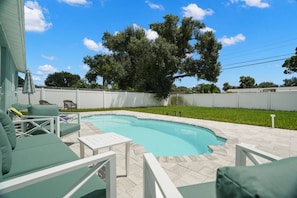 A swimming pool with a curved edge, surrounded by a white fence, and a grassy area with a large tree. - Modern 3BR Pool Home · 15 Min to Beaches (Pinellas Park)