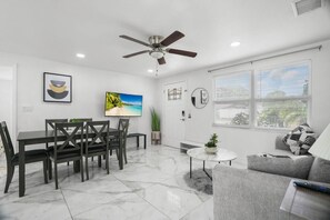 A modern living room with a dining area, a ceiling fan, a TV, and a large window with blinds. - Modern 3BR Pool Home · 15 Min to Beaches (Pinellas Park)