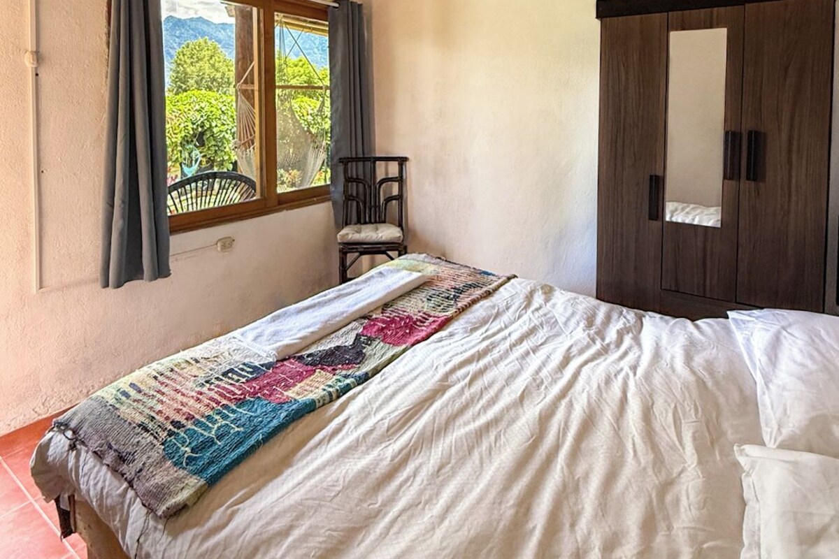 A bedroom with a wooden ceiling, a bed with a patterned blanket, a window with curtains, and a wardrobe.