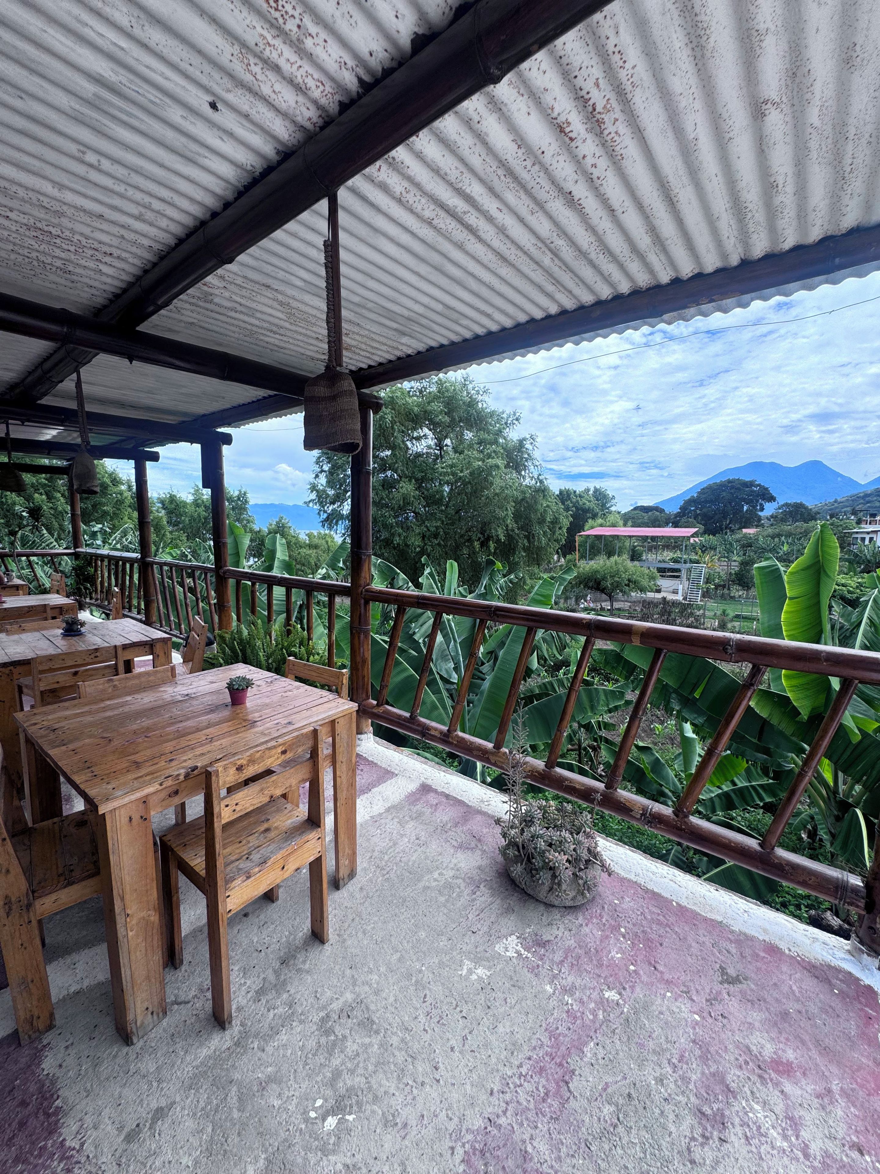An outdoor seating area with wooden tables and chairs, a metal railing, and a view of a mountainous landscape.