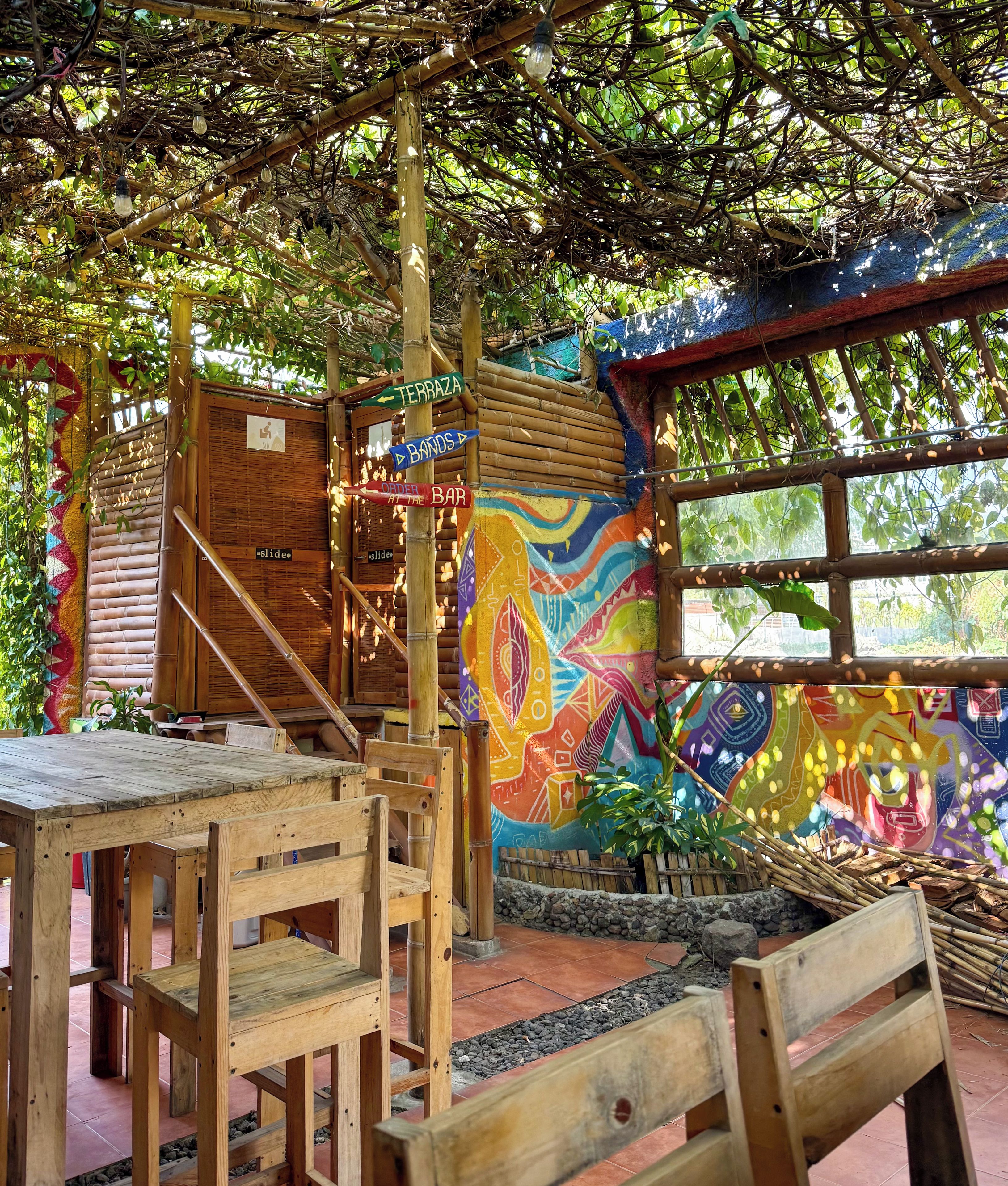 A rustic outdoor seating area with wooden tables and chairs, a bamboo staircase, and a colorful mural on the wall.