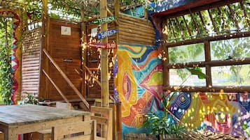 Un área de estar al aire libre de estilo rústico, con mesas y sillas de madera, una escalera de bambú y un mural colorido en la pared.