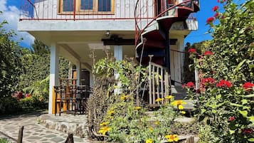 Un edificio de dos pisos con un balcón, una escalera y un jardín con diversas plantas y flores.
