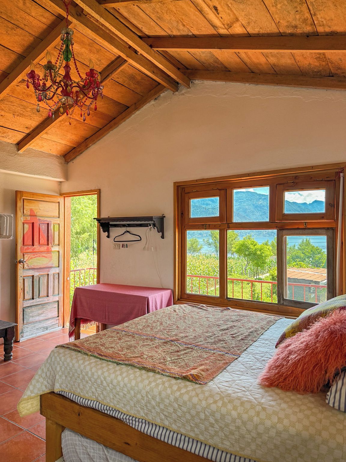 A bedroom with a wooden ceiling, a bed with a patterned bedspread, a window with a view of mountains, and a door leading outside.