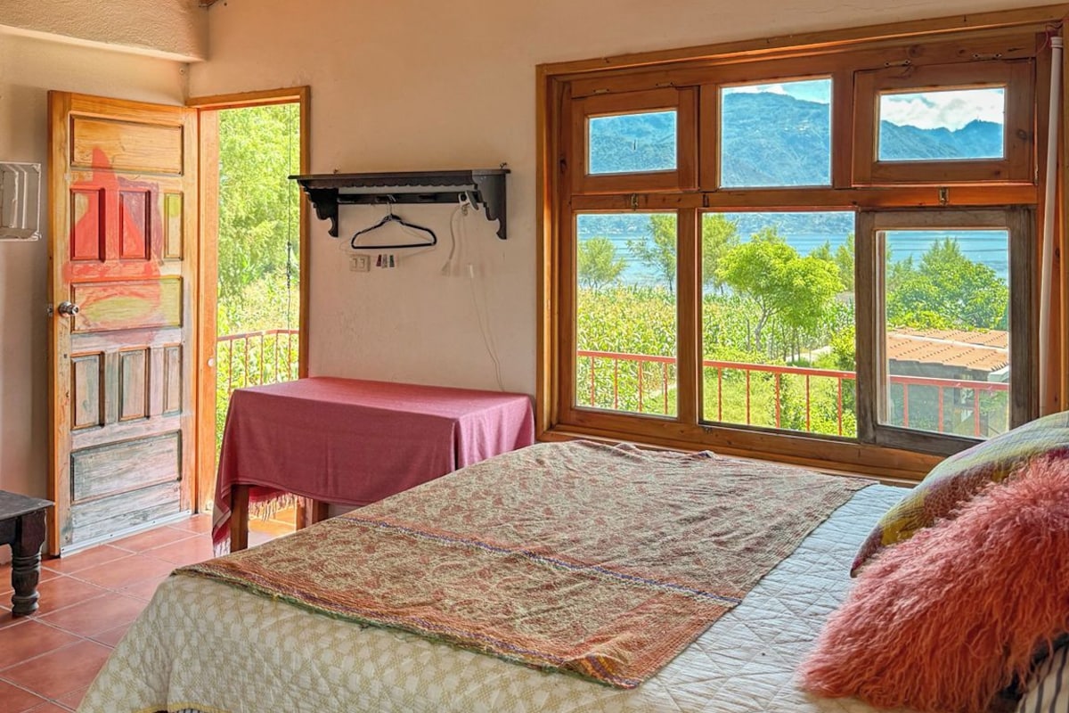 A bedroom with a wooden ceiling, a bed with a patterned bedspread, a window with a view of mountains, and a door leading outside.
