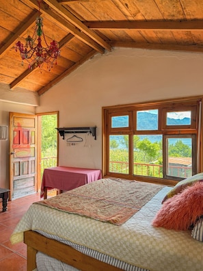 A bedroom with a wooden ceiling, a bed with a patterned bedspread, a window with a view of mountains, and a door leading outside. - Luna y Sol Atitlan (San Pedro La Laguna)