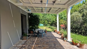 En overdækket terrasse med stenbelægning, et bord, stole og plantekrukker.